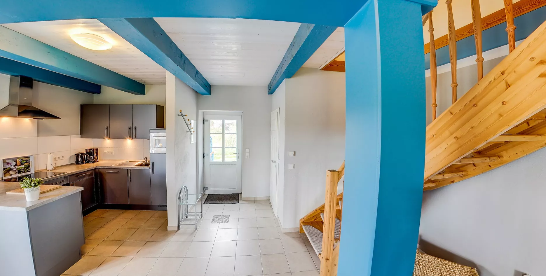 Offene Küche und Eingangsbereich mit dunklen Fronten, Holztreppe und blauen Deckenbalken.